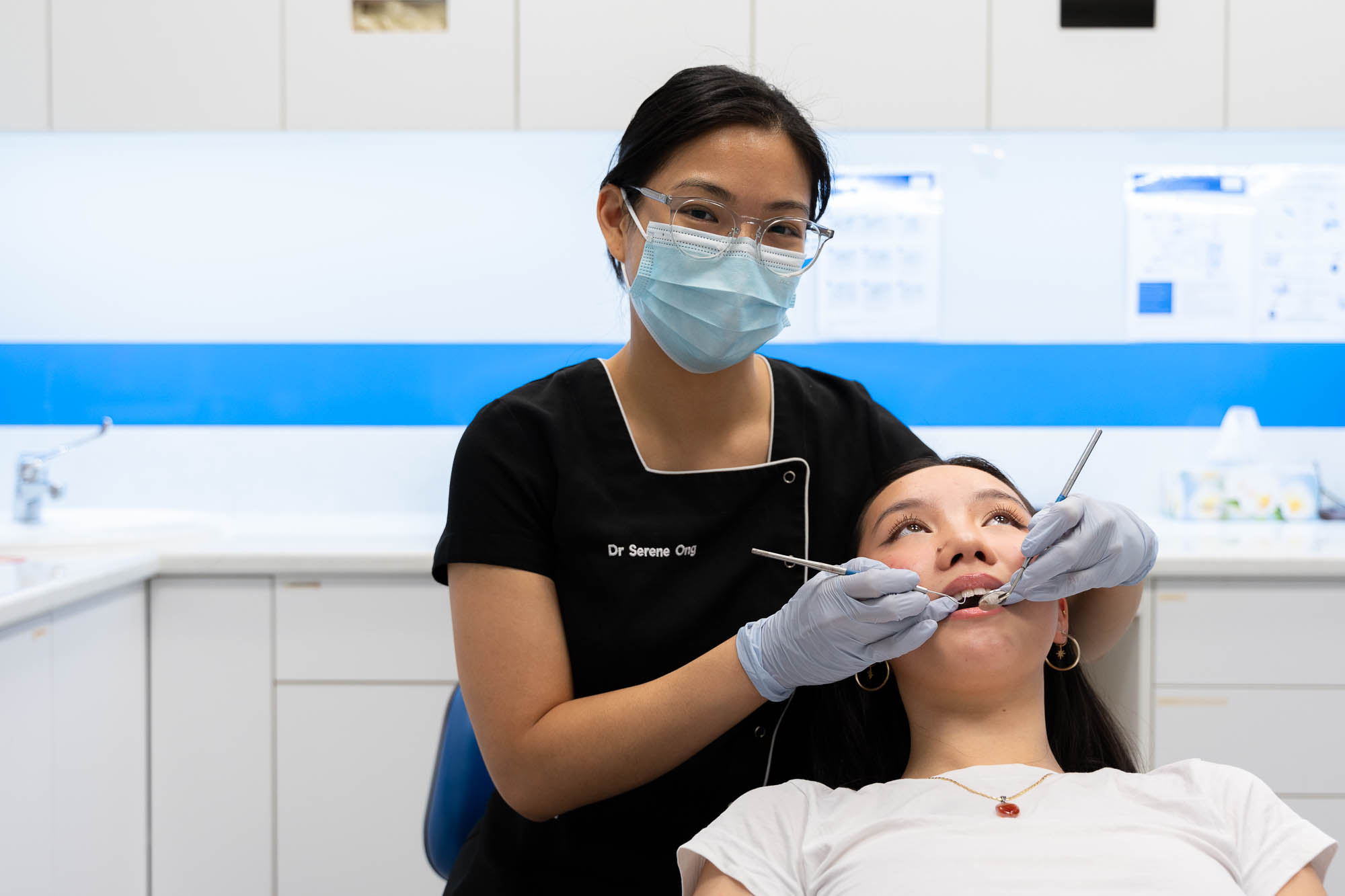 DR Serene Ong in maske looking at camera while assisting detnal patient in chair.
